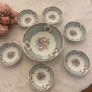 Może przedstawiać: Zestaw siedmiu porcelanowych misek. Duża miska i sześć mniejszych misek mają jasnoniebieską obwódkę ze złotymi zdobieniami. Każda miska ma pośrodku kwiatowy wzór z różowymi, niebieskimi i fioletowymi różami.