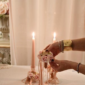 May include: Two lit pink candles with floral decorations and a small floral arrangement. A person's hands hold a small floral bouquet with ribbons. The background is a soft pink curtain.