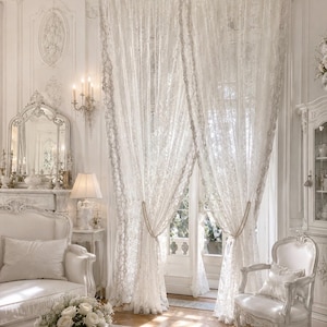 May include: A pair of floor-length white lace curtains featuring an intricate floral pattern. The curtains are gathered at the centre with beaded tiebacks, allowing light to filter through the delicate fabric while covering a large window and door.