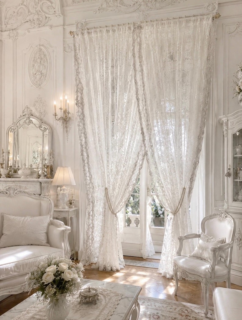May include: A pair of floor-length white lace curtains featuring an intricate floral pattern. The curtains are gathered at the centre with beaded tiebacks, allowing light to filter through the delicate fabric while covering a large window and door.