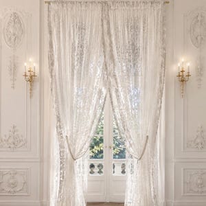 May include: A pair of floor-length white lace curtains featuring an intricate floral pattern and scalloped edges. The sheer fabric is gathered and held back by beaded tiebacks, hanging from a gold-toned rod over a set of double glass doors.
