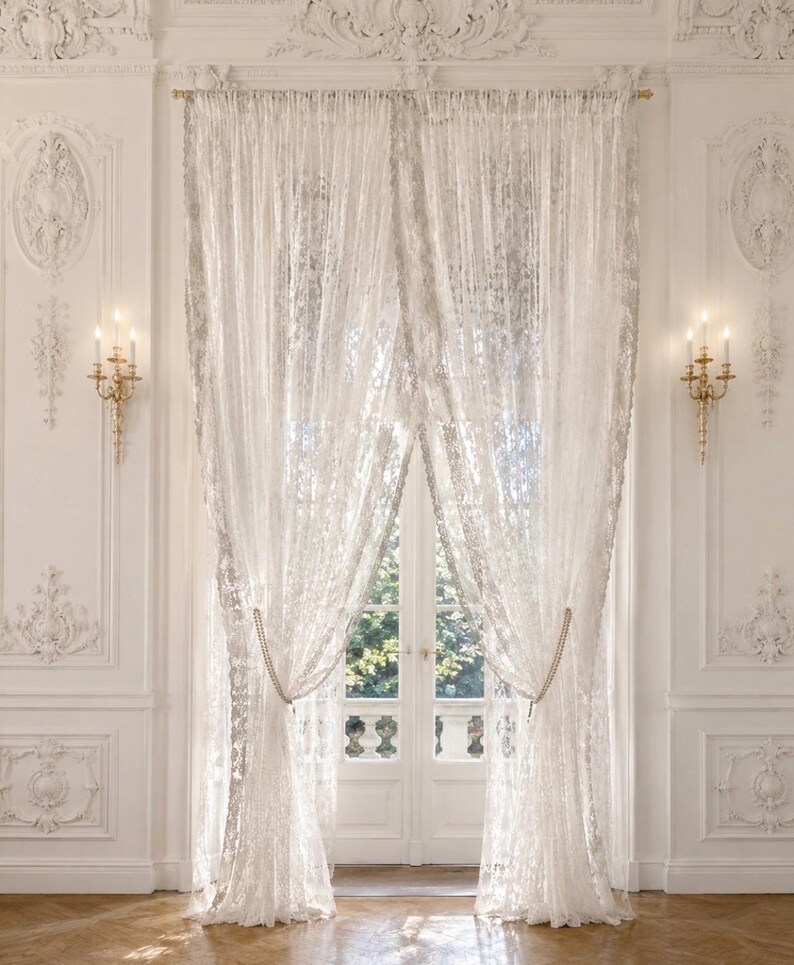 May include: A pair of floor-length white lace curtains featuring an intricate floral pattern and scalloped edges. The sheer fabric is gathered and held back by beaded tiebacks, hanging from a gold-toned rod over a set of double glass doors.