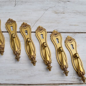 Könnte beinhalten: Sechs antike Messing-Türgriffe mit verzierten Details und Schlüssellöchern. Die Griffe sind in einer Reihe auf einer weißen Holzoberfläche angeordnet.