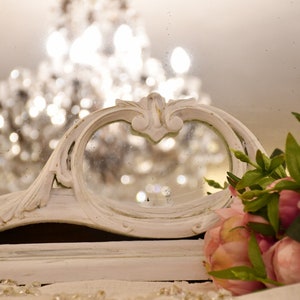 May include: A white ornate mirror with a decorative frame and a reflection of a chandelier. Pink flowers are in the foreground.