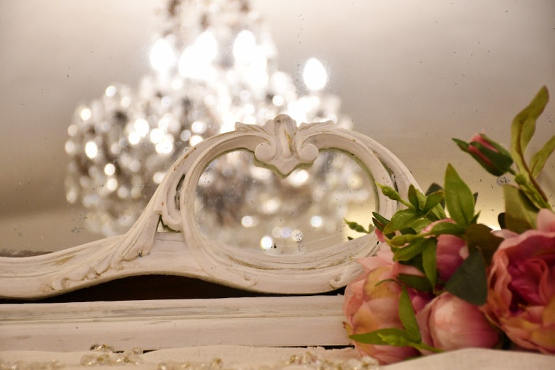 May include: A white ornate mirror with a decorative frame and a reflection of a chandelier. Pink flowers are in the foreground.