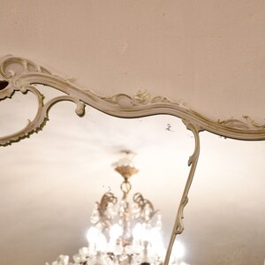 May include: A white ornate mirror with a decorative, scrolled frame. The mirror reflects a chandelier with crystal accents.