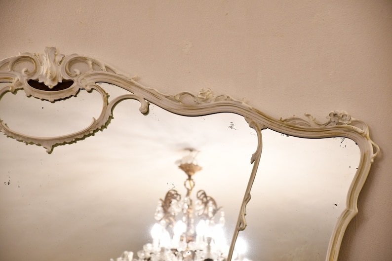 May include: A white ornate mirror with a decorative, scrolled frame. The mirror reflects a chandelier with crystal accents.