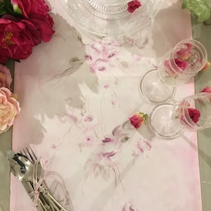 Può includere: Un runner da tavolo floreale rosa con un delicato disegno floreale. Il runner è decorato con rose rosa e ha uno sfondo rosa chiaro. Ci sono due bicchieri di vetro trasparente con rose rosa all'interno, un piatto di vetro trasparente e un set di posate d'argento.