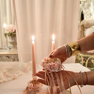 May include: A delicate scene with two lit pink candles and a floral arrangement. A person holds a small floral ring with ribbons. The setting is soft and feminine, with a lace tablecloth and a vase of flowers in the background.