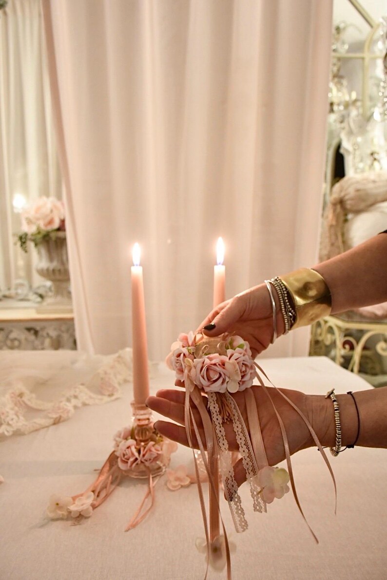 May include: A delicate scene with two lit pink candles and a floral arrangement. A person holds a small floral ring with ribbons. The setting is soft and feminine, with a lace tablecloth and a vase of flowers in the background.