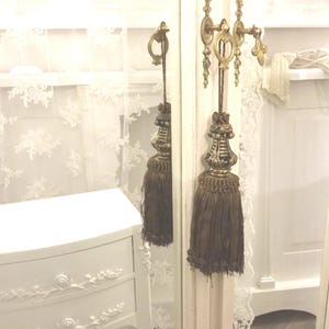 May include: A white vanity mirror with a decorative gold tassel hanging from the top. The mirror has a white frame with ornate details.