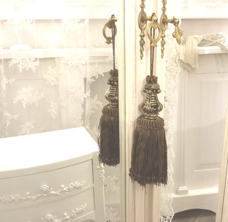 May include: A white vanity mirror with a decorative gold tassel hanging from the top. The mirror has a white frame with ornate details.