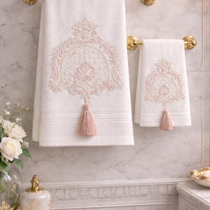 Può includere: Due asciugamani bianchi con intricati ricami rosa e nappe, appesi a portasciugamani dorati. Gli asciugamani presentano un design ornato, aggiungendo un tocco di eleganza a un ambiente bagno.