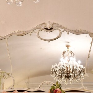 May include: A large, ornate, white, antique mirror with a decorative, carved frame. The mirror is reflecting a crystal chandelier with clear glass crystals.