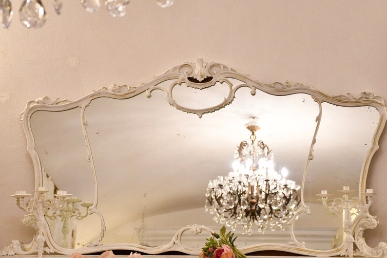 May include: A large, ornate, white, antique mirror with a decorative, carved frame. The mirror is reflecting a crystal chandelier with clear glass crystals.