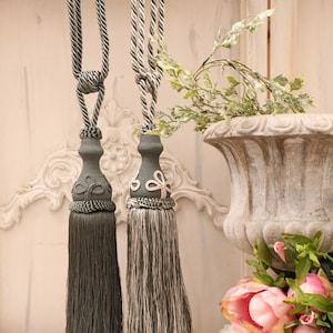May include: Two decorative tassel tiebacks, one in gray and one in black and white, with a floral design on the top. The tassels are hanging from a white wall with a vase of flowers in the background.
