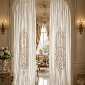 May include: Elegant cream-colored curtains with ornate, embroidered embellishments hang in an arched doorway. The curtains frame a view of a luxurious living room with a chandelier, sofa, and a table with flowers. The room is decorated in neutral tones.