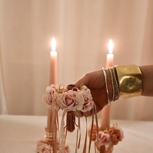 May include: A small bouquet of pink roses with lace and ribbon is held in a hand, with two lit pink candles behind it. A gold cuff bracelet and multiple silver bracelets are also visible. The image has a romantic, decorative theme.