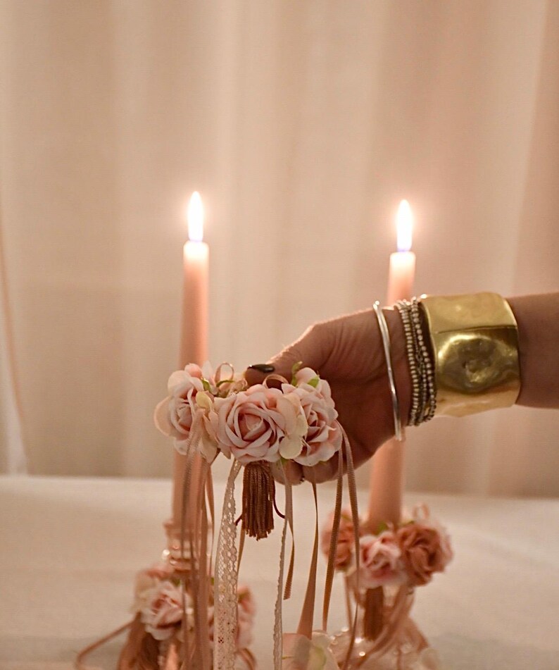 May include: A small bouquet of pink roses with lace and ribbon is held in a hand, with two lit pink candles behind it. A gold cuff bracelet and multiple silver bracelets are also visible. The image has a romantic, decorative theme.