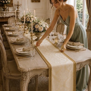 Puede incluir: Elegante mesa de comedor con un camino de mesa color crema con ribete dorado. La mesa está decorada con fina porcelana, copas de cristal y centros de mesa florales. La habitación tiene una estética clásica y lujosa.