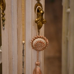 Peut inclure: Une porte de cabinet en bois blanc avec une poignée en métal doré et un pompon. Le pompon est fait d'un tissu beige avec un motif rose et blanc.