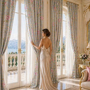 Può includere: Una persona in un abito di raso color champagne è in piedi vicino a una finestra, tenendo una tenda floreale. La stanza presenta finiture dorate ornate, un lampadario e tende azzurre con motivi floreali rosa. La vista esterna comprende un balcone e uno specchio d'acqua.