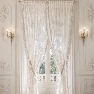 May include: A pair of floor-length white lace curtains hanging from a gold curtain rod. The curtains feature an intricate floral pattern and are gathered at the sides with beaded tiebacks, allowing light to filter through the delicate fabric.
