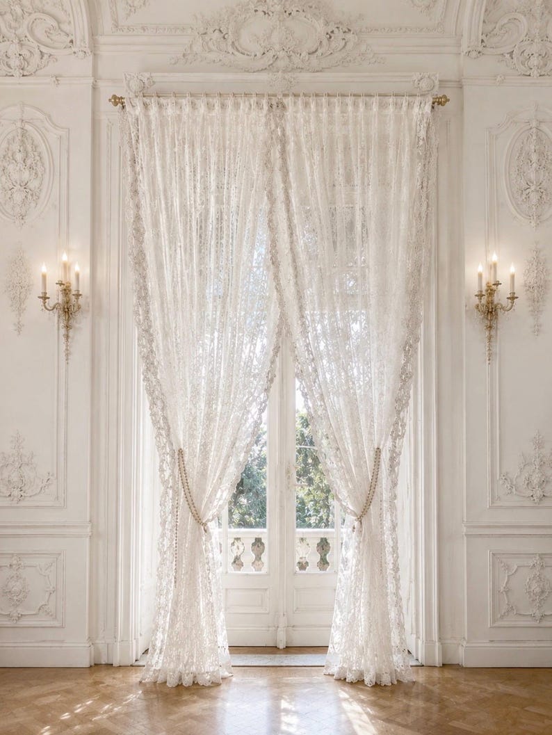 May include: A pair of floor-length white lace curtains hanging from a gold curtain rod. The curtains feature an intricate floral pattern and are gathered at the sides with beaded tiebacks, allowing light to filter through the delicate fabric.