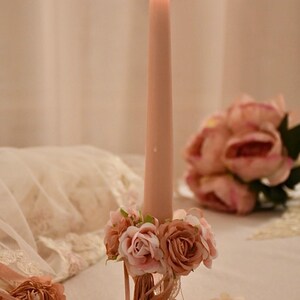 May include: A lit, pink taper candle in a decorative glass holder decorated with pink and peach roses. The candle is central, with a soft, blurred background featuring more floral arrangements.