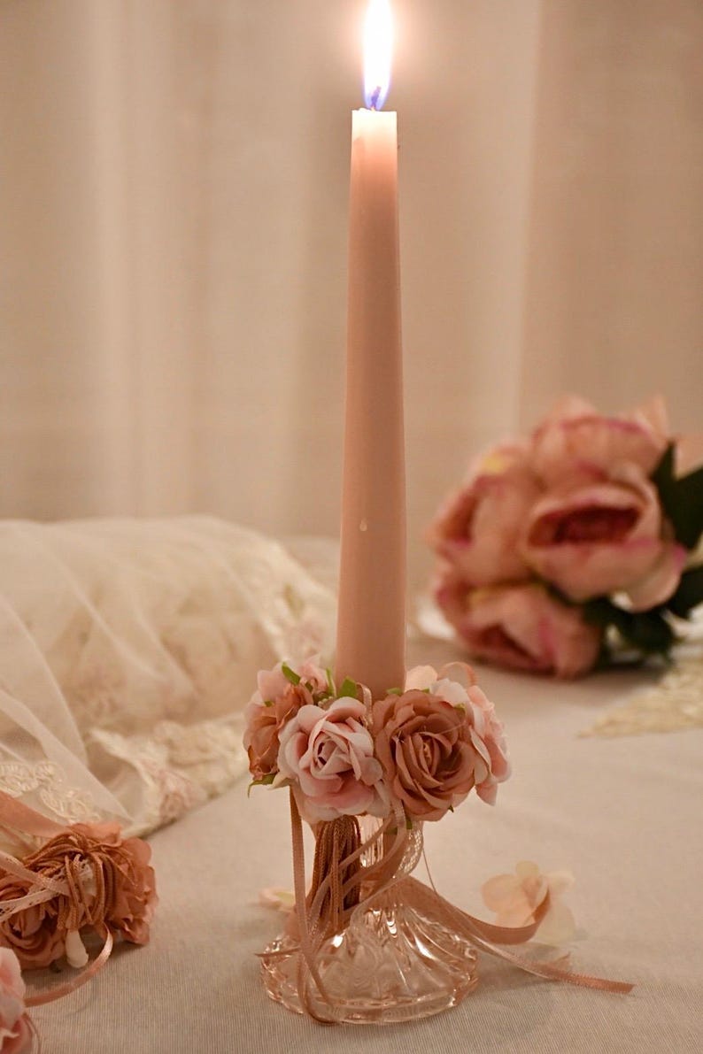 May include: A lit, pink taper candle in a decorative glass holder decorated with pink and peach roses. The candle is central, with a soft, blurred background featuring more floral arrangements.