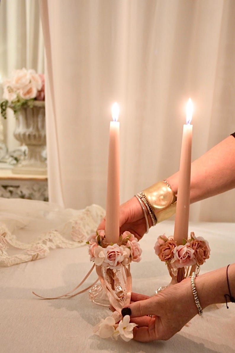 May include: Two lit, pale pink taper candles in ornate glass holders, decorated with pink and peach roses. The holders are being held by hands, with a gold cuff bracelet visible. The scene is set against a soft, neutral background.