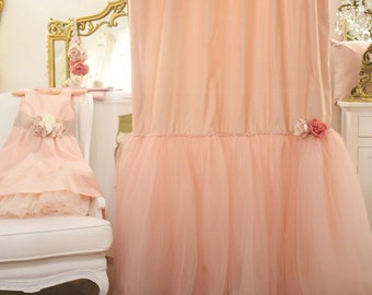 May include: A pale pink tulle curtain with a ruffled hem and a floral embellishment. The curtain is hanging in a room with a white chair and a white dresser.