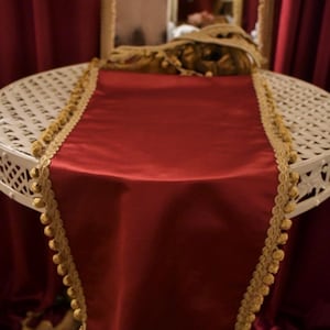 May include: A red satin table runner with a gold trim featuring small, round embellishments. The runner is draped over a white, ornate, round table. The background includes red curtains and a mirror.