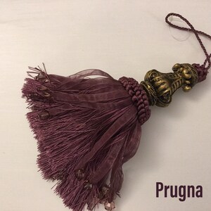 May include: A deep purple tassel with a decorative gold metal top. The tassel is made of a soft, silky fabric and has a long, flowing fringe. The word "Prugna" is printed below the tassel.