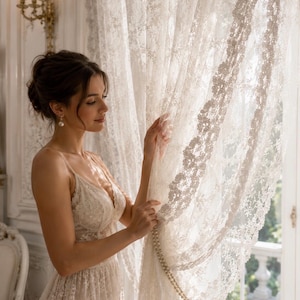 May include: A floor-length ivory lace dress featuring a V-neckline, thin spaghetti straps, and intricate floral embroidery throughout the fabric. The dress is paired with a matching sheer lace curtain panel adorned with a delicate pearl strand.