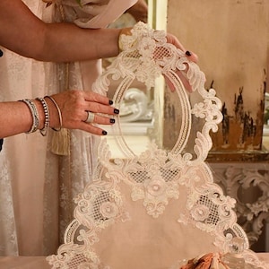 Può includere: Una tovaglia in pizzo color crema con un motivo floreale. La tovaglia è presentata, mostrando i suoi intricati dettagli e la sua delicata consistenza. Lo sfondo include una tenda e elementi decorativi.