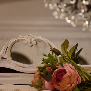 May include: A white ornate mirror with a distressed finish, featuring a heart-shaped opening. A bouquet of pink roses and green leaves is placed in front of the mirror.