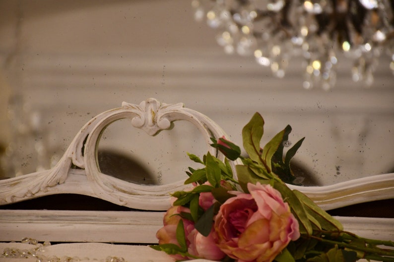 May include: A white ornate mirror with a distressed finish, featuring a heart-shaped opening. A bouquet of pink roses and green leaves is placed in front of the mirror.