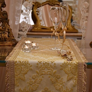 Può includere: Un runner da tavolo in damasco dorato e bianco con bordo a frangia. Due flute da champagne con accenti di nastro dorato sono sul runner, insieme a tre piccoli portacandele di vetro.