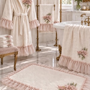 Peut inclure: Un ensemble de salle de bain couleur crème comprenant un peignoir à capuche, des serviettes et un tapis de bain rectangulaire. Chaque pièce présente une bordure à volants rose pâle et une application florale décorative dans les tons rose, blanc et vert.