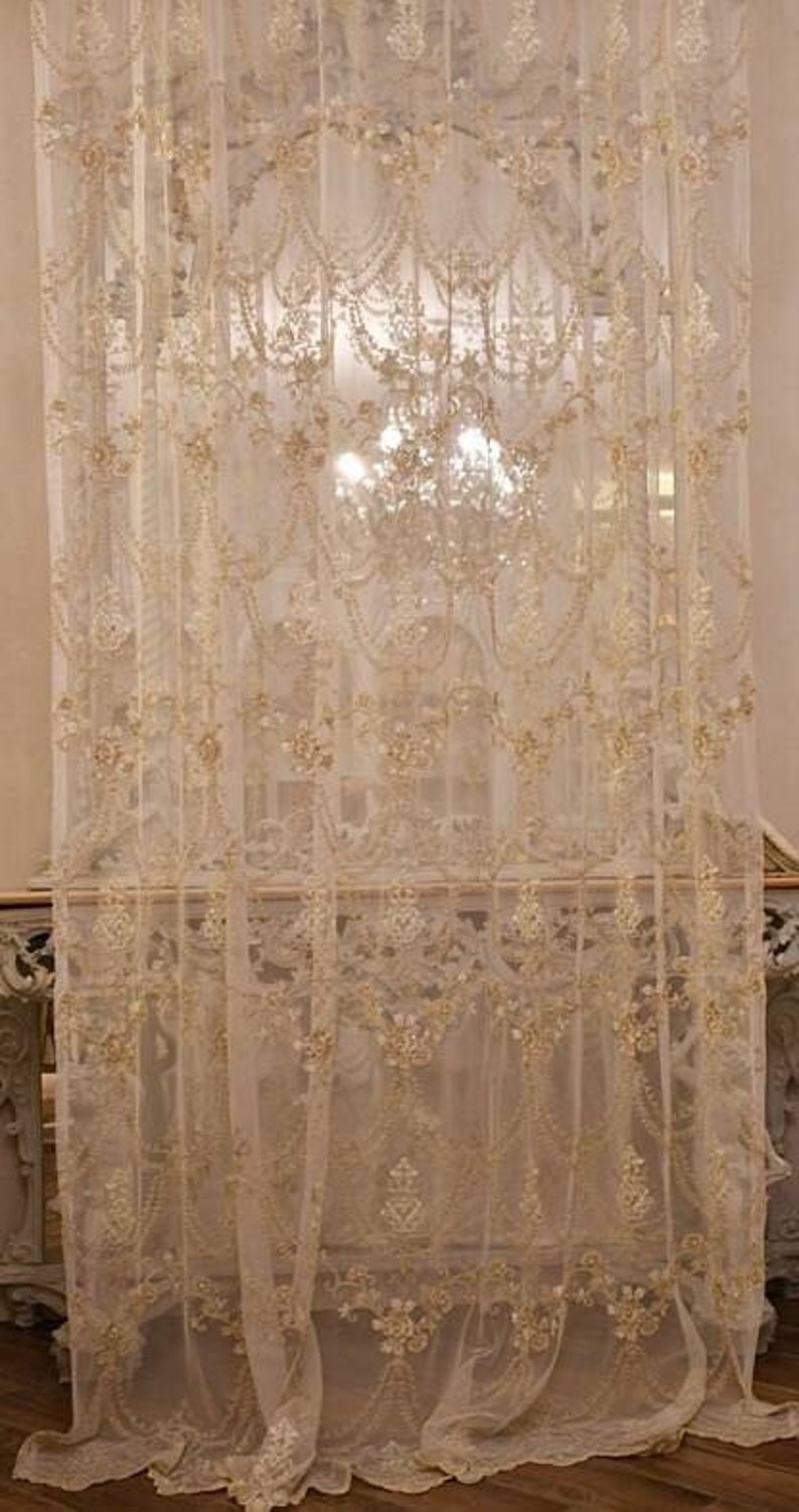 May include: White sheer curtains with a delicate floral pattern and gold embroidery. The curtains are hung in a room with a large window and a chandelier.
