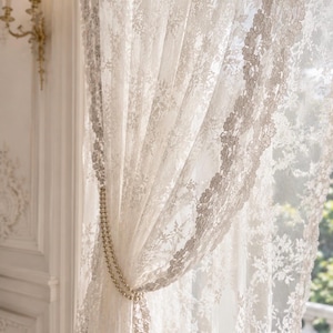 May include: A sheer white lace curtain featuring an intricate floral pattern and scalloped edges. The curtain is gathered to one side and secured with a double-strand pearl tieback, allowing light to filter through the delicate, semi-transparent fabric.