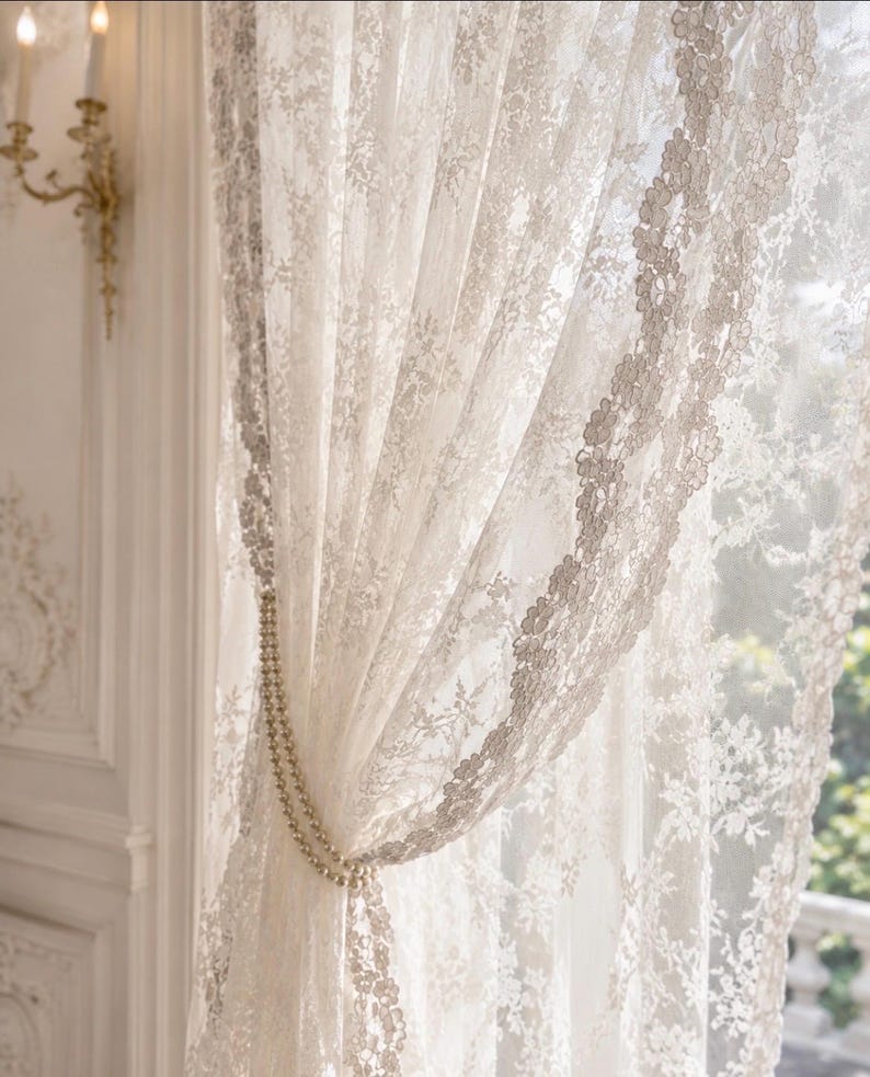 May include: A sheer white lace curtain featuring an intricate floral pattern and scalloped edges. The curtain is gathered to one side and secured with a double-strand pearl tieback, allowing light to filter through the delicate, semi-transparent fabric.