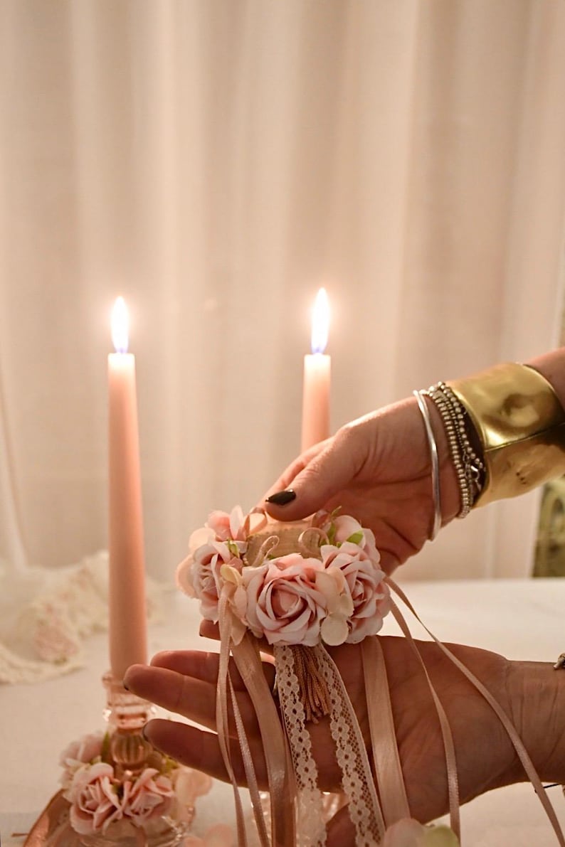 May include: A floral wrist corsage with pink roses, ribbons, and lace is held in front of two lit pink candles. The corsage is accented with a small tassel. A gold cuff bracelet and silver bracelets adorn the wrist.