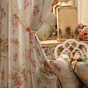 Könnte beinhalten: Hellblaue Blumenvorhänge mit rosa Rosen und grünen Blättern. Die Vorhänge werden mit einem dekorativen Spitzenvorhanghalter mit einem rosa Rosenakzent zurückgehalten. Passende Kissen und ein weißer Stuhl vervollständigen die romantische Einrichtung.