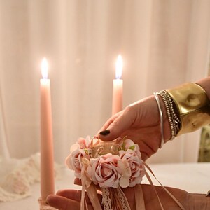 May include: A floral wrist corsage with pink roses, ribbons, and lace is held in front of two lit pink candles. The corsage is accented with a small tassel. A gold cuff bracelet and silver bracelets adorn the wrist.