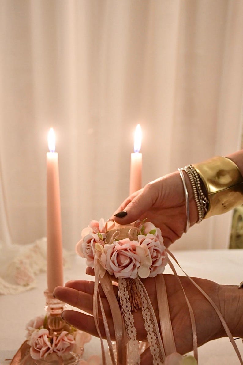 May include: A floral wrist corsage with pink roses, ribbons, and lace is held in front of two lit pink candles. The corsage is accented with a small tassel. A gold cuff bracelet and silver bracelets adorn the wrist.
