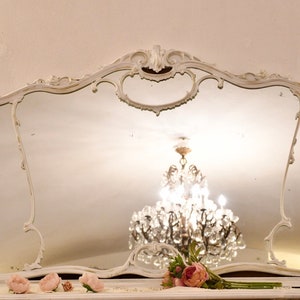 May include: A large, white, ornate mirror with a decorative, scalloped frame. The mirror reflects a crystal chandelier hanging from the ceiling.