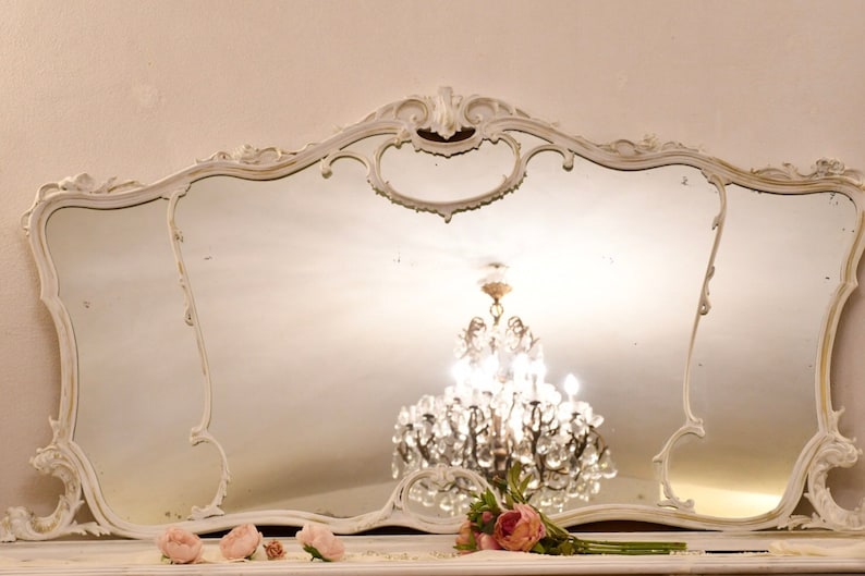 May include: A large, white, ornate mirror with a decorative, scalloped frame. The mirror reflects a crystal chandelier hanging from the ceiling.
