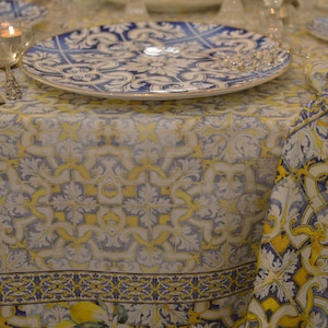 May include: A white tablecloth with a blue and yellow tile pattern. The tablecloth has a border of lemons and leaves. There are two plates and silverware on the table.
